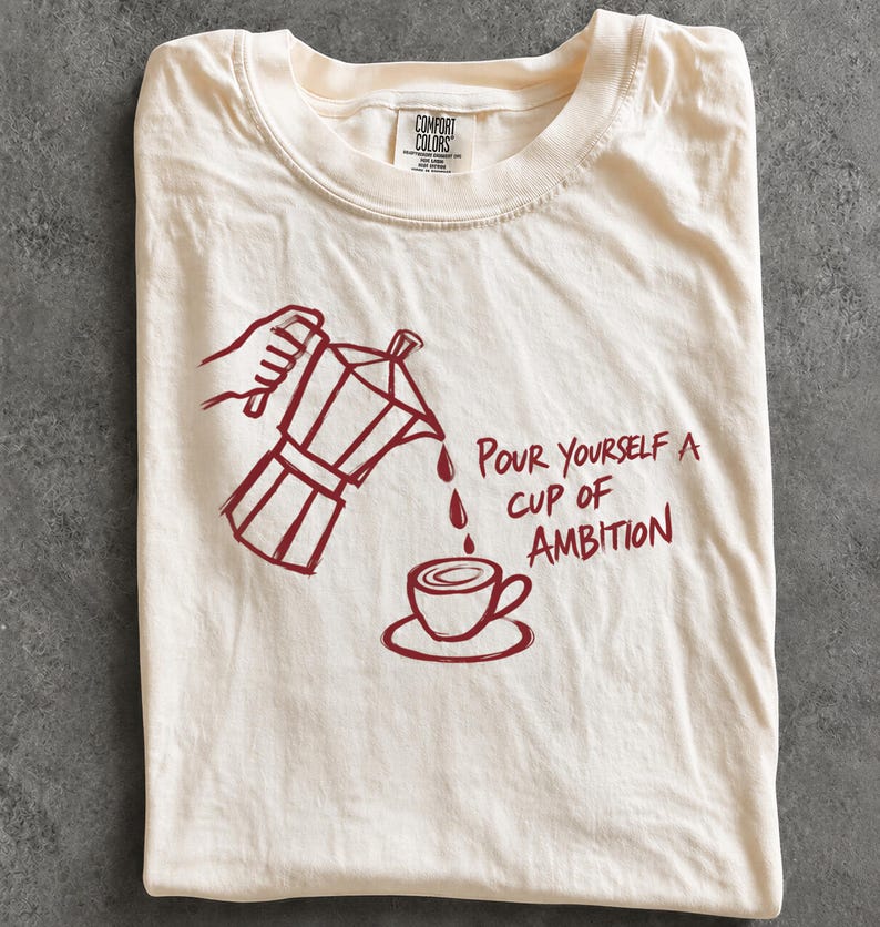 A white t-shirt with a red graphic design depicting a coffee pot pouring coffee into a cup, accompanied by the text "Pour yourself a cup of ambition".