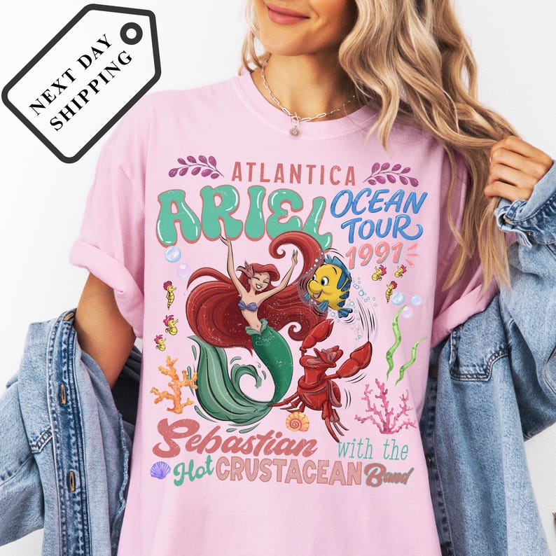 A woman wearing a pink t-shirt with a graphic design featuring a mermaid and a crab, along with text promoting the Ariel Ocean Tour.