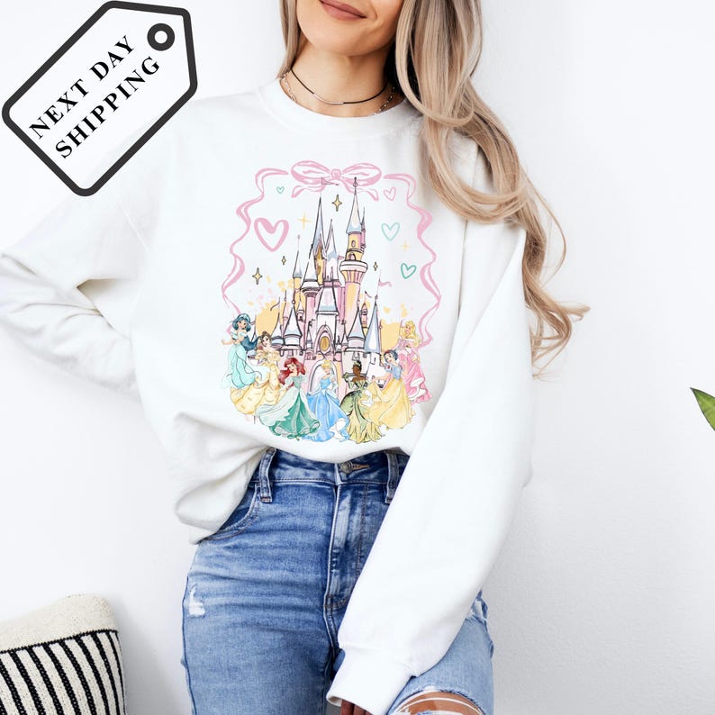 A woman wearing a white sweatshirt with a Disney princess castle design and a pink ribbon around it.