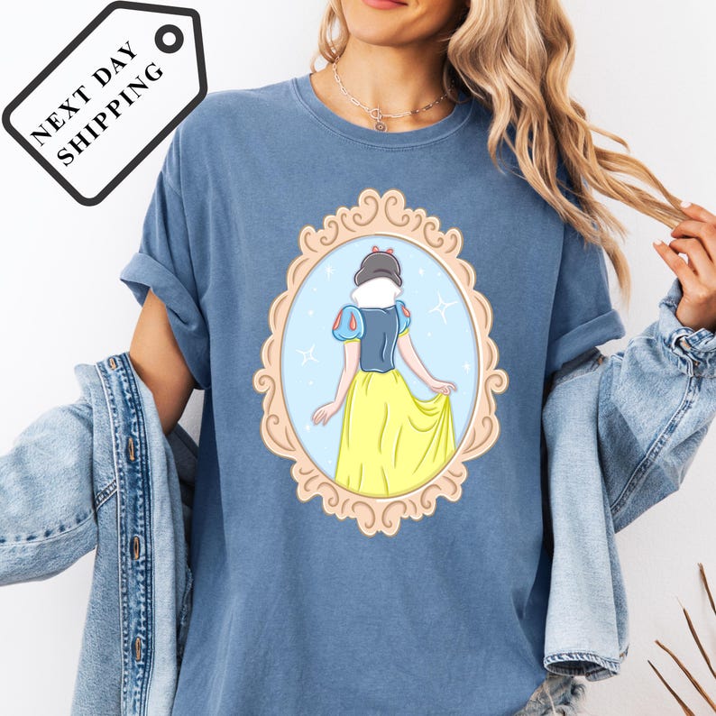 A woman wearing a blue t-shirt with a graphic of a princess in a yellow dress, standing in a decorative frame.