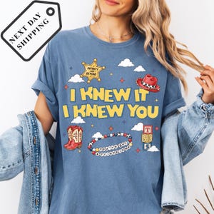 A woman wearing a blue t-shirt with a graphic design featuring a cowboy hat, a star, and various other cartoon-style elements.