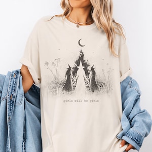 A woman wearing a white t-shirt with a graphic design of two horses and a crescent moon, along with the text "girls will be girls".