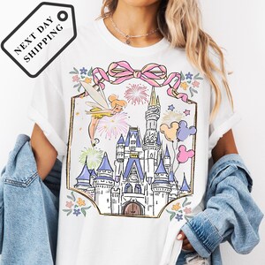 A woman wearing a white t-shirt with a colorful illustration of a castle and various Disney characters, including Tinker Bell and Mickey Mouse, on a white background.