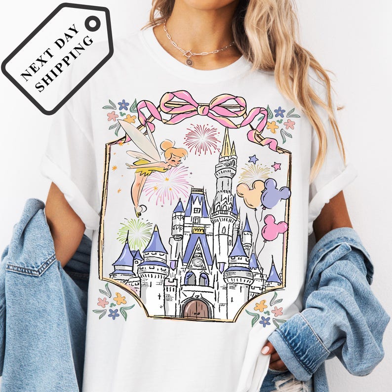 A woman wearing a white t-shirt with a colorful illustration of a castle and various Disney characters, including Tinker Bell and Mickey Mouse, on a white background.