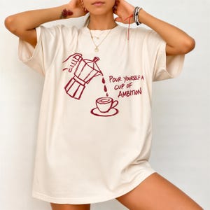 A woman wearing a white t-shirt with a graphic design of a coffee pot pouring coffee into a cup, accompanied by the text "Pour yourself a cup of ambition".