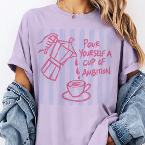 A person wearing a purple t-shirt with a graphic design depicting a coffee cup pouring from a coffee pot, accompanied by the text "Pour Yourself a Cup of Ambition".