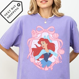 A woman wearing a purple t-shirt with a Disney Princess design, including a pink ribbon, flowers, and a castle.
