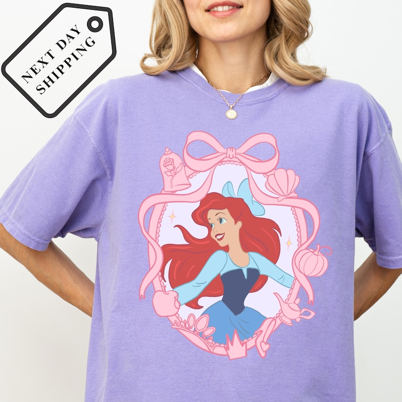 A woman wearing a purple t-shirt with a Disney Princess design, including a pink ribbon, flowers, and a castle.