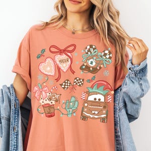 A woman wearing a pink t-shirt with a Christmas-themed design, including a Santa Claus hat, a mug, and other festive elements.