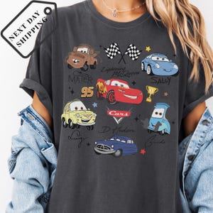 A person wearing a gray t-shirt with various cartoon car characters printed on it, including Lightning McQueen, Sally, and other iconic vehicles from the Cars movie franchise.