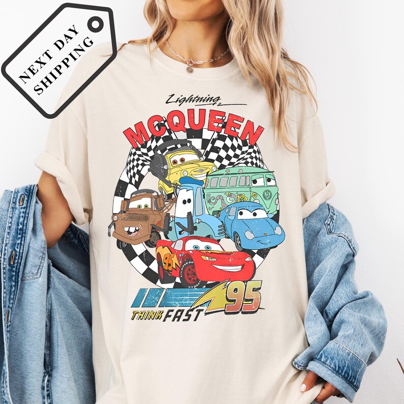 A person wearing a white t-shirt with a graphic of various cartoon cars from the Cars movie franchise, including Lightning McQueen, speeding around a checkered track.