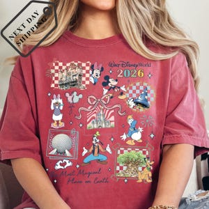 A person wearing a red t-shirt with various Disney characters and landmarks printed on it, including Mickey Mouse, Minnie Mouse, Goofy, and the Walt Disney World theme park.