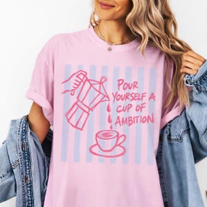 A woman wearing a pink t-shirt with a graphic design of a coffee cup and the text "Pour yourself a cup of ambition" printed on it.