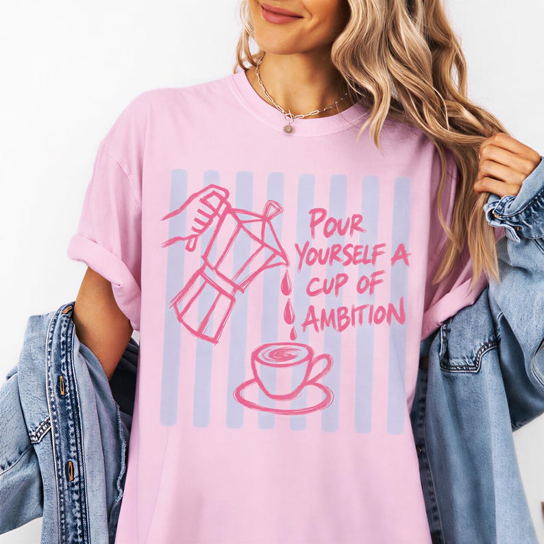 A woman wearing a pink t-shirt with a graphic design of a coffee cup and the text "Pour yourself a cup of ambition" printed on it.