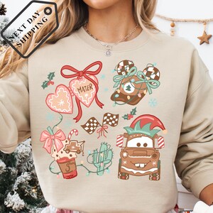 A person wearing a beige sweater with a Christmas-themed design featuring various holiday-related elements such as hearts, a Santa hat, a mug, and a car.