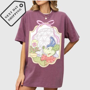 A woman wearing a purple t-shirt with a graphic design featuring a cartoon character and various decorative elements.