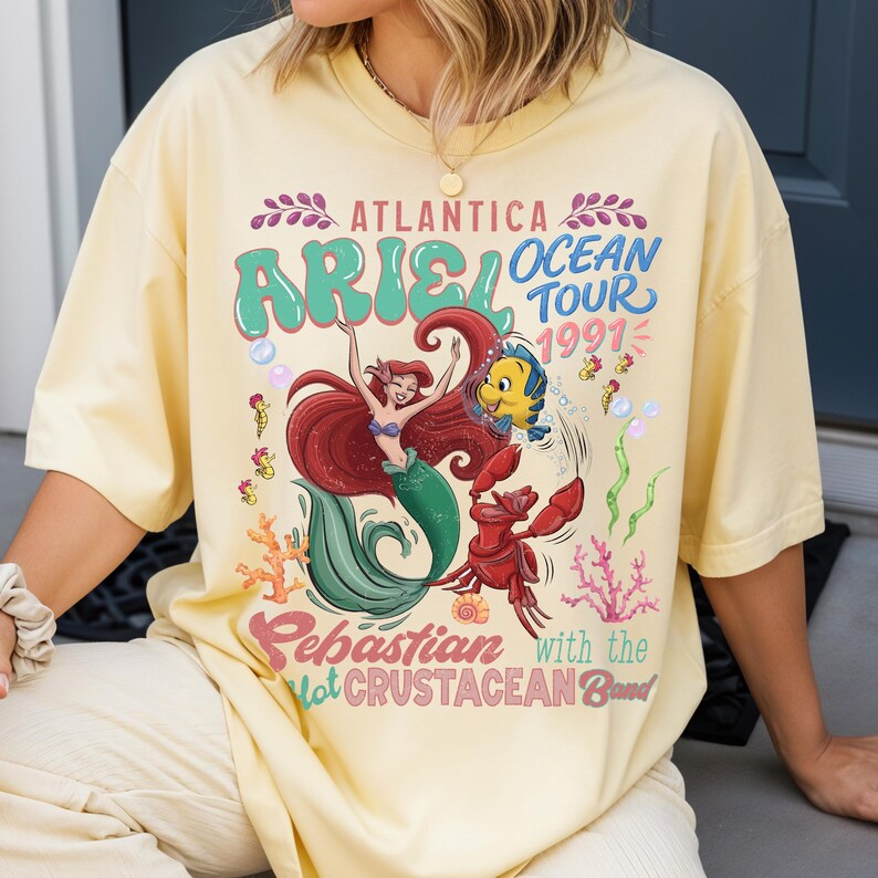 A person wearing a yellow t-shirt with a colorful graphic design featuring the mermaid Ariel from the Disney film "The Little Mermaid".