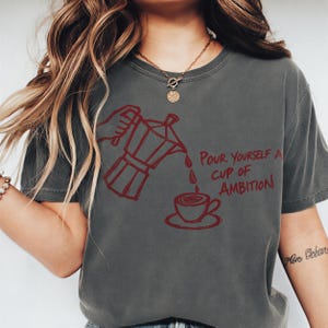 A woman wearing a gray t-shirt with a graphic design of a coffee pot pouring coffee into a cup, accompanied by the text "Pour Yourself a Cup of Ambition".