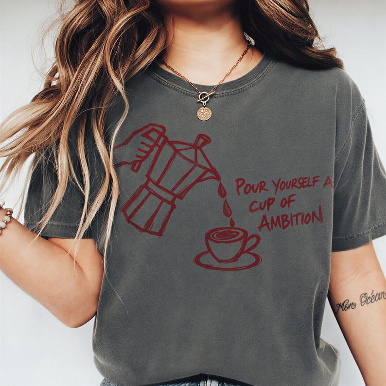 A woman wearing a gray t-shirt with a graphic design of a coffee pot pouring coffee into a cup, accompanied by the text "Pour Yourself a Cup of Ambition".