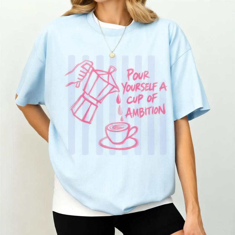 A woman wearing a light blue t-shirt with a graphic design of a coffee cup and a hand pouring coffee.