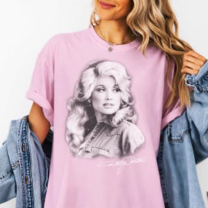 A woman wearing a pink t-shirt with a black and white portrait of a woman printed on it, along with a denim jacket.