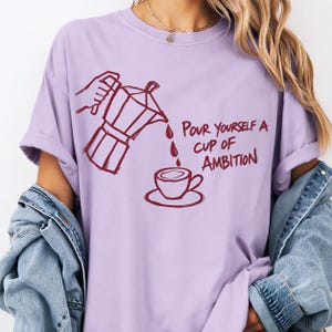 A person wearing a purple t-shirt with a graphic design depicting a coffee pot pouring coffee into a cup, accompanied by the text "Pour yourself a cup of ambition".