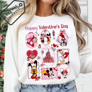 A white long-sleeved shirt with a graphic design featuring various Disney characters and elements, including Minnie Mouse, Mickey Mouse, Donald Duck, Goofy, and other iconic figures. The design also includes a castle, hearts, and other festive symbols associated with Valentine's Day.