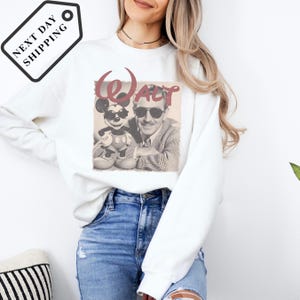 A woman wearing a white sweatshirt with a graphic of Mickey Mouse and Walt Disney on it, along with a tag that says "Next day shipping".