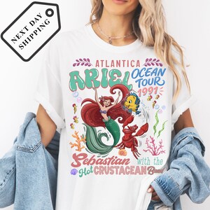 A white t-shirt with a colorful graphic design featuring a mermaid character and text related to the Ariel Ocean Tour.