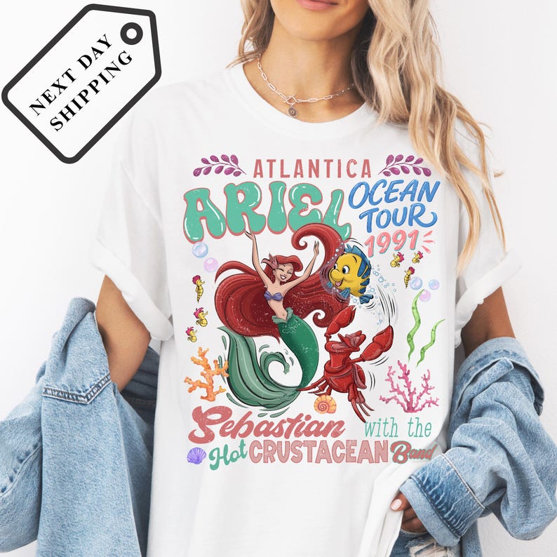 A white t-shirt with a colorful graphic design featuring a mermaid character and text related to the Ariel Ocean Tour.