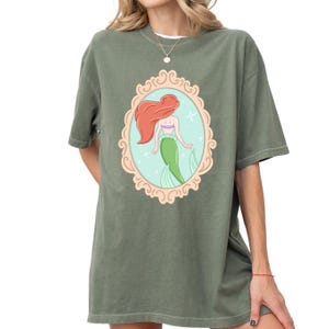 A woman wearing a green t-shirt with a graphic of a mermaid in a frame.