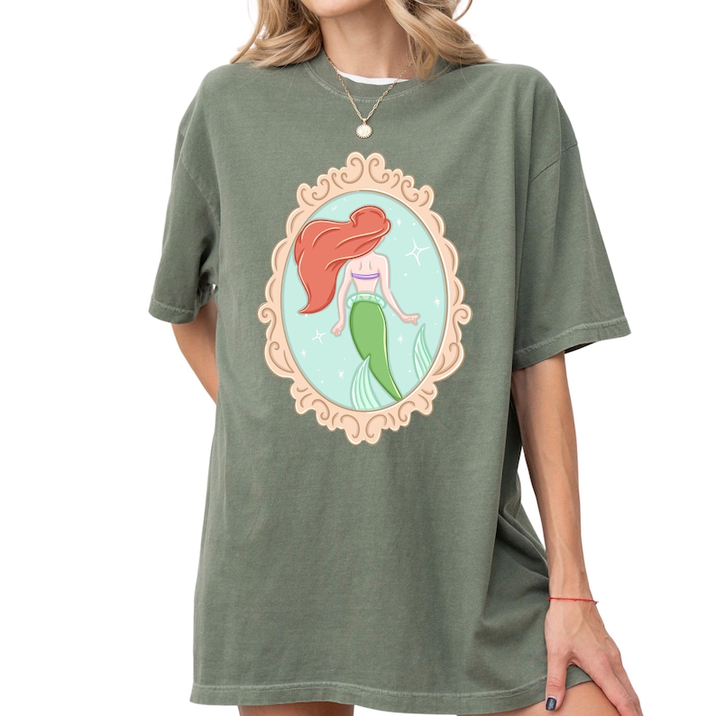 A woman wearing a green t-shirt with a graphic of a mermaid in a frame.