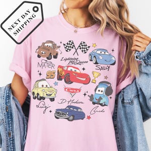 A person wearing a pink t-shirt with various cartoon car characters and text related to the Cars movie franchise.