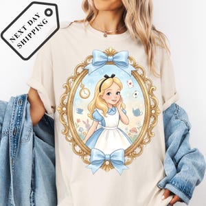 A woman wearing a white t-shirt with a graphic of a young girl in a blue dress and a clock, surrounded by a decorative frame.