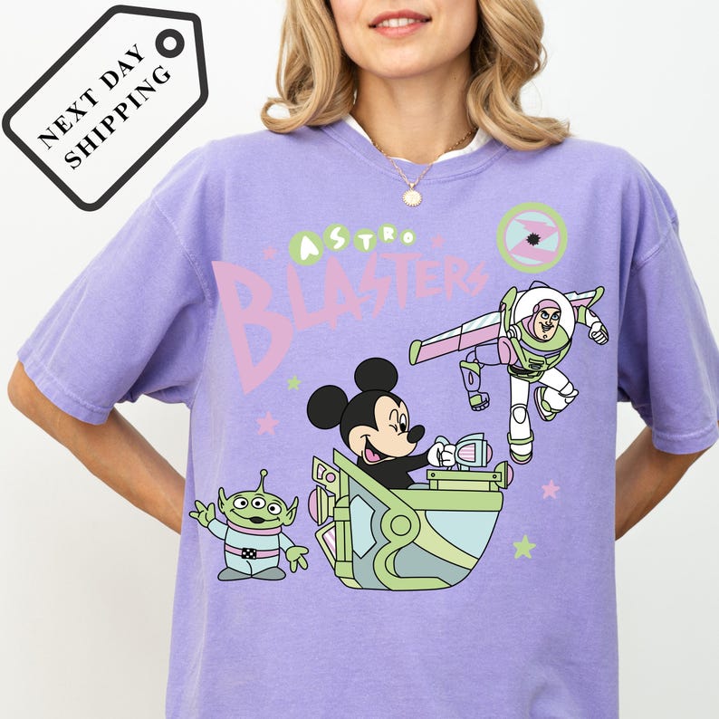 Pu&ograve; includere: Una maglietta lilla con una grafica di Mickey Mouse, un alieno e Buzz Lightyear con la scritta "Astro Blasters". Il design include accenti rosa, verdi e bianchi. La maglietta ha una vestibilit&agrave; ampia.