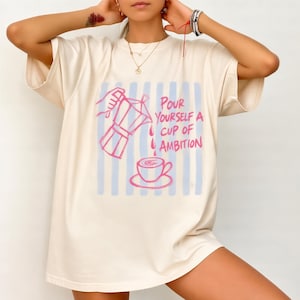 A woman wearing a white t-shirt with a graphic design of a coffee pot pouring coffee and the text "Pour yourself a cup of ambition" printed on it.
