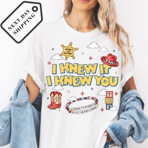 A woman wearing a white t-shirt with a graphic design featuring a cowboy hat, clouds, and stars, along with the text "I KNEW IT I KNEW YOU".