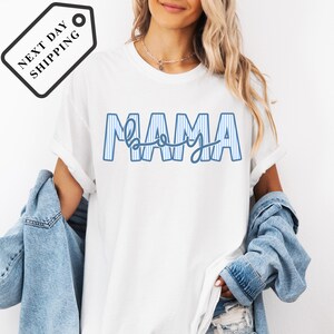 A woman wearing a white t-shirt with the word "MAMA" printed on it, along with a denim jacket.