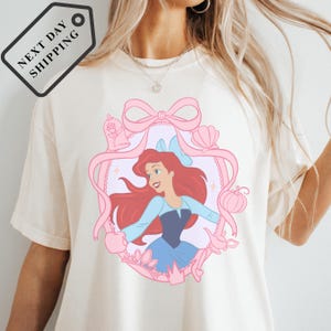 A white t-shirt with a pink and blue illustration of a young girl with red hair, wearing a blue dress and surrounded by various Disney-themed elements.