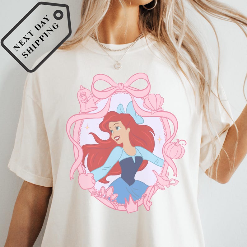 A white t-shirt with a pink and blue illustration of a young girl with red hair, wearing a blue dress and surrounded by various Disney-themed elements.