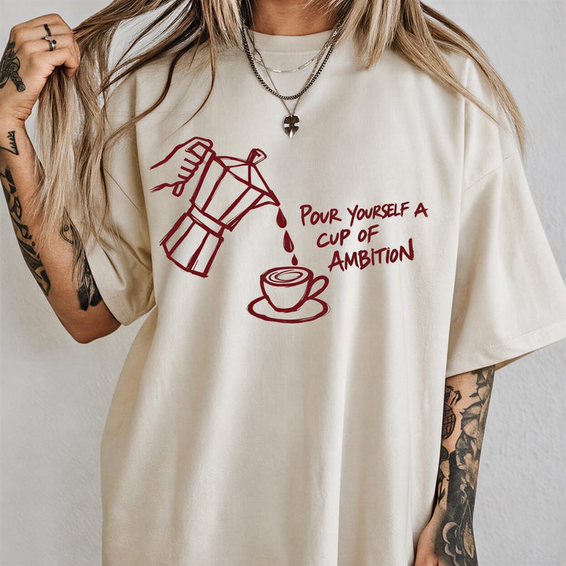 A person wearing a white t-shirt with a graphic design of a coffee pot pouring coffee into a cup, accompanied by the text "Pour yourself a cup of ambition".