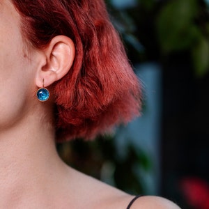 May include: A close-up of a person's ear with a blue and gold earring. The earring is a small, round, blue stone with a gold setting. The person has red hair.
