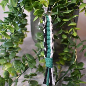 May include: A green, black and white braided keyring with a silver key ring. The keyring has a tassel at the bottom.