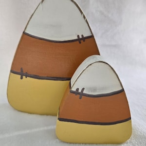 May include: Two wooden candy corn decorations. The larger piece is behind the smaller one. Each has a white top, a brown middle, and a yellow bottom. Black lines and cross-shaped details separate the colors. The decorations are on a white surface.