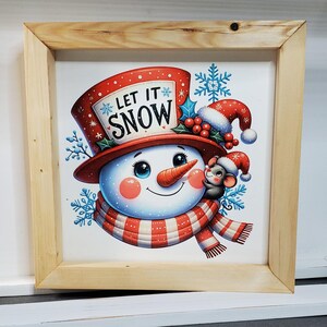 May include: A framed Christmas-themed print featuring a snowman with a carrot nose, wearing a red hat and scarf. The text "LET IT SNOW" is displayed on the hat. A small mouse wearing a Santa hat is next to the snowman. The frame is made of light-colored wood.