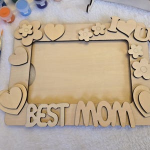 May include: A wooden photo frame with the words "BEST MOM" and "I ❤️ U" cut out. The frame is decorated with heart and flower shapes. Paintbrushes and paint pots are visible.