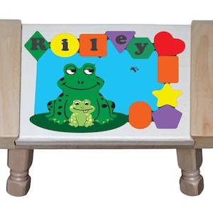 May include: A wooden children's puzzle table with a frog theme. The puzzle features the name "Riley" spelled out with colorful geometric shapes. The table has a light wood frame and legs, and a white surface with a blue background.