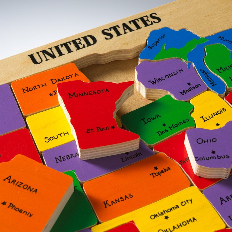 Puede incluir: Rompecabezas de mapa de los Estados Unidos de madera con piezas de estados coloridas. La pieza roja de Minnesota est&aacute; levantada, con la capital, St. Paul, etiquetada. Otros estados como Arizona, Kansas e Iowa son visibles.