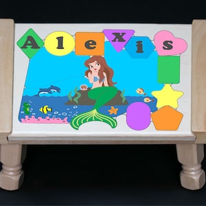 May include: A wooden stool with a colorful puzzle top featuring a mermaid, fish, and the name "Alexis". The puzzle pieces are shaped like different geometric forms.