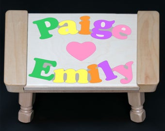 Puede incluir: Un taburete de madera con un corazón y los nombres Paige y Emily escritos en letras de colores. Las letras están recortadas en la madera y son de diferentes colores, incluyendo verde, amarillo, naranja, morado y rosa.